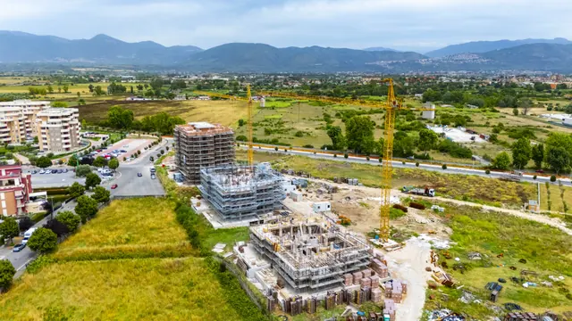 Il cantiere dal drone: Edificio A in completamento, Edificio B in elevazione, Edificio C alle prime strutture — fotografia di cantiere, giugno 2026